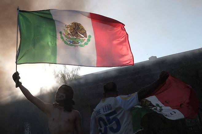 8일(현지시각) 미국 캘리포니아주 로스앤젤레스에서 이민자 단속에 저항하는 시위대가 불탄 차량이 피워올리는 연기 속에서 멕시코 국기를 흔들고 있다. 사진 AFP 연합뉴스
