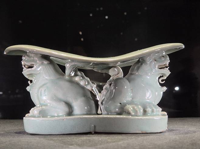 청자사자모양베개. 머리를 대는 넓은 판 아래 목에 방울을 단 사자 두 마리가 입을 벌리고 엉덩이를 맞대고 웅크려 앉아 있다.