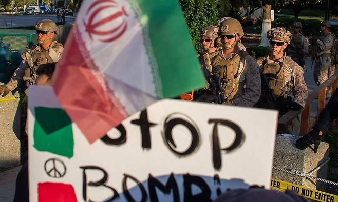 18일 미국 캘리포니아주 로스앤젤레스의 윌셔 연방건물 앞을 미국 해병대가 지키는 가운데 “이란 전쟁 반대” 집회에 참석한 시민들이 ‘폭격을 멈춰라’는 등의 팻말을 들고 있다. AFP 연합뉴스