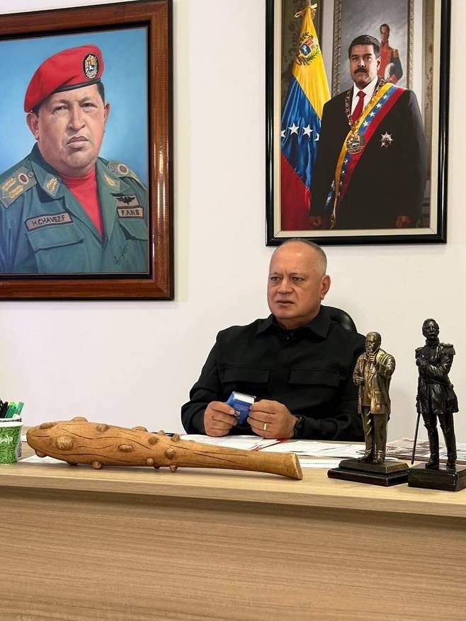 7일 디오스다도 카베요 베네수엘라 내무장관이 손에 헌법을 들고 집무실 책상에 앉아 있다. 책상 위에는 카베요 장관이 진행하는 TV쇼 '망치로 때리기'의 상징인 망방이가 놓여 있고, 뒤쪽 벽에는 본인의 정치적 뿌리인 우고 차베스 전 베네수엘라 대통령의 초상화(왼쪽)와 지난 3일 미국에 체포된 니콜라스 마두로 베네수엘라 대통령의 초상화가 걸려 있다. /'콘엘마조단도(망치로 때리기)' 방송 공식 인스타그램