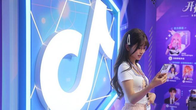 한 여성이 화려한 틱톡 로고 앞에 서서 스마트폰을 보고 있다