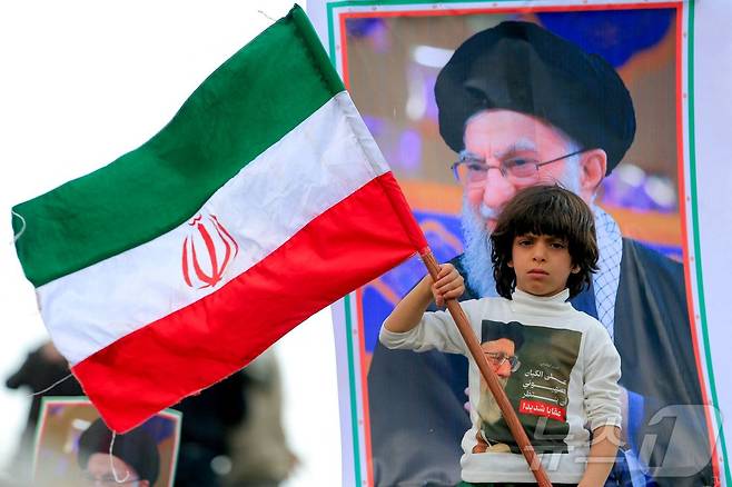 이란 국기를 든 소년. 뒤에는 미국의 공습을 받고 사망한 이란 전 최고 지도자 아야톨라 알리 하메네이. ⓒ AFP=뉴스1