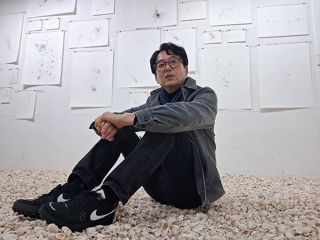 경기 수원 실험공간 유즈에서 근작전 ‘잔여의 정치학’을 펼치고 있는 김재남 작가를 지난 14일 전시장에서 만났다. 고향인 전남 여수에서 가져와 전시장 바닥에 가득 깔아놓은 꼬막 껍데기 위에 작가가 앉았다. 그 뒤로 목탄 가루를 뿌려 별이나 성운처럼 확산되는 이미지를 만든 평면 드로잉 작업들이 벽에 내걸려 있다. 노형석 기자