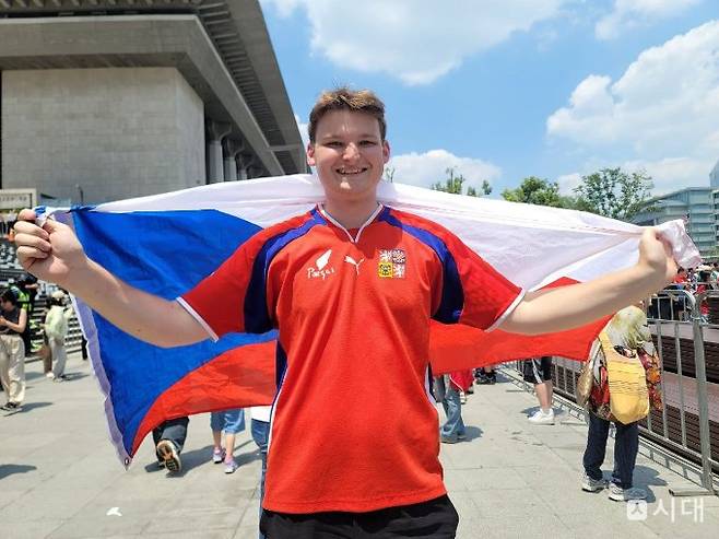 12일 오전 '2026 국제축구연맹(FIFA) 북중미 월드컵' 한국-체코전 경기를 응원하기 위해 광화문 광장을 찾은 체코인 웨이따(21)씨가 양팔을 벌려 체코 국기를 두른 채 미소를 짓고 있다. /사진=조아현 기자