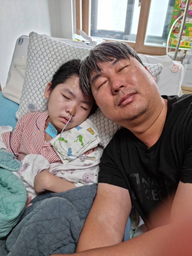 ▲고된 현장 일을 마치고 돌아와 아들 곁에 기대어 짧은 쪽잠을 청하는 아빠 김정훈 씨. 새는 빗물을 막아내는 단단한 아빠는, 가장 가까운 곳에서 가족의 눈물을 닦아주는 든든한 버팀목이다. ⓒ프레시안
